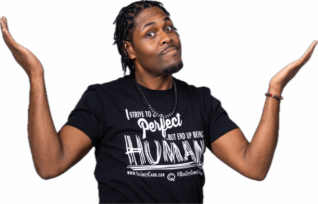 A man with braided hair gestures with open hands, wearing a black t-shirt that says, "I strive to be perfect but end up being HUMAN."