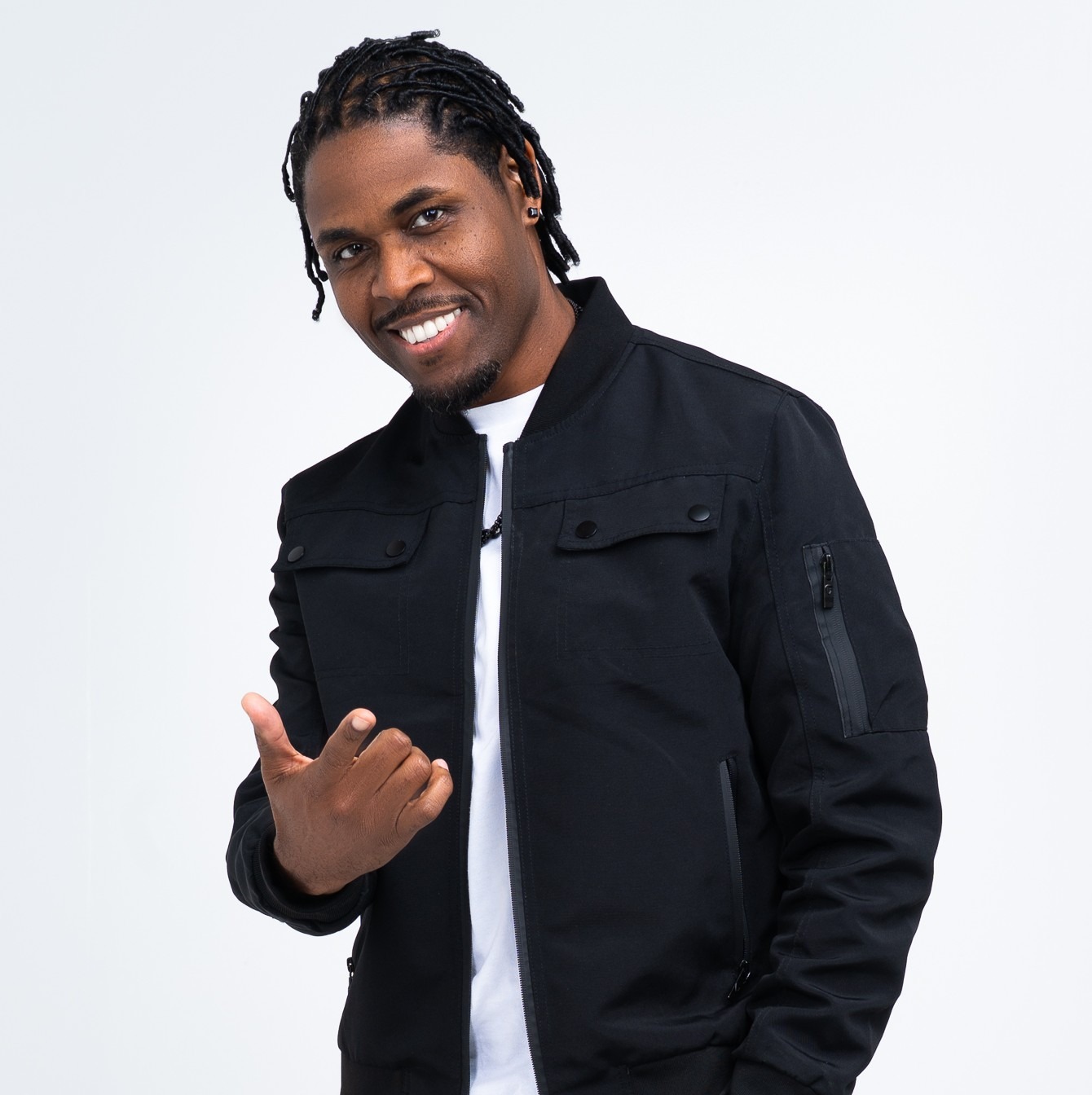 Smiling man in a black jacket and white t-shirt gives a thumbs-up gesture, suggesting positivity and confidence against a plain white background.