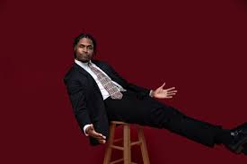 A well-dressed man in a suit sits on a stool, leaning back playfully against a deep red background. His confident pose and open gestures convey charisma.