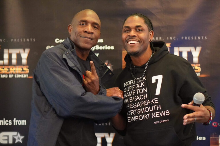 Two men pose together smiling and gesturing, with one wearing a black hoodie and the other a jacket. A promotional backdrop for a comedy series is visible behind them.
