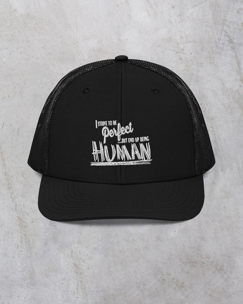 Black trucker cap featuring the phrase "I strive to be perfect but end up being HUMAN" in white text. Emphasizes a relatable message about imperfections.