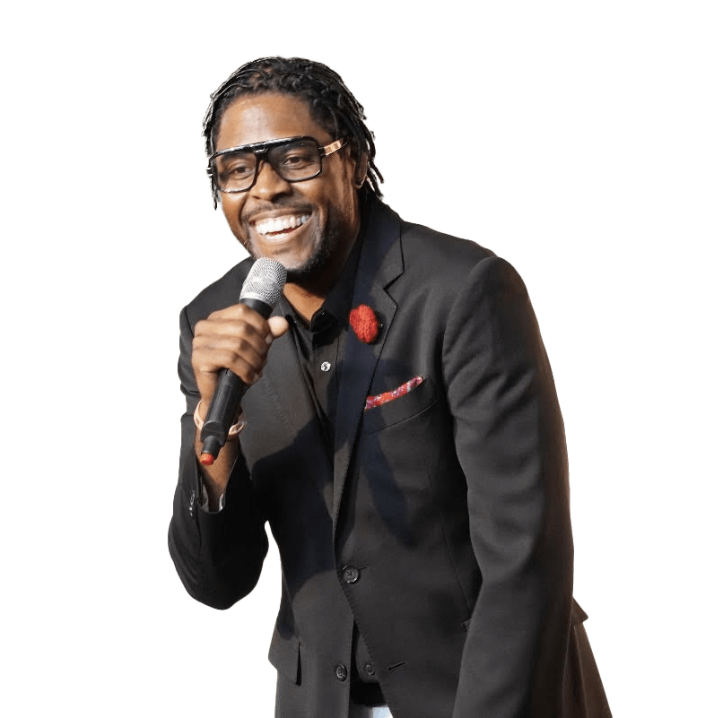 Smiling performer in a black suit holds a microphone, exuding charisma on stage. His look, featuring stylish glasses and a flower pin, enhances his engaging presence.